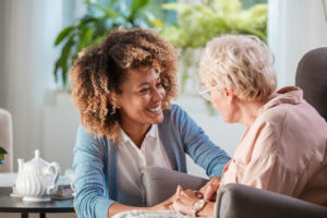 Smiling young female caregiver holds hands and talks with senior woman in living room