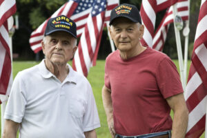 One male Veteran wearing Korea-Vietnam Veteran hat and another mature man wearing Vietnam Veteran hat in front of American flags outside