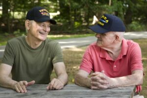 Two male Veterans wearing Veteran hats talking to one another on a bench in nature